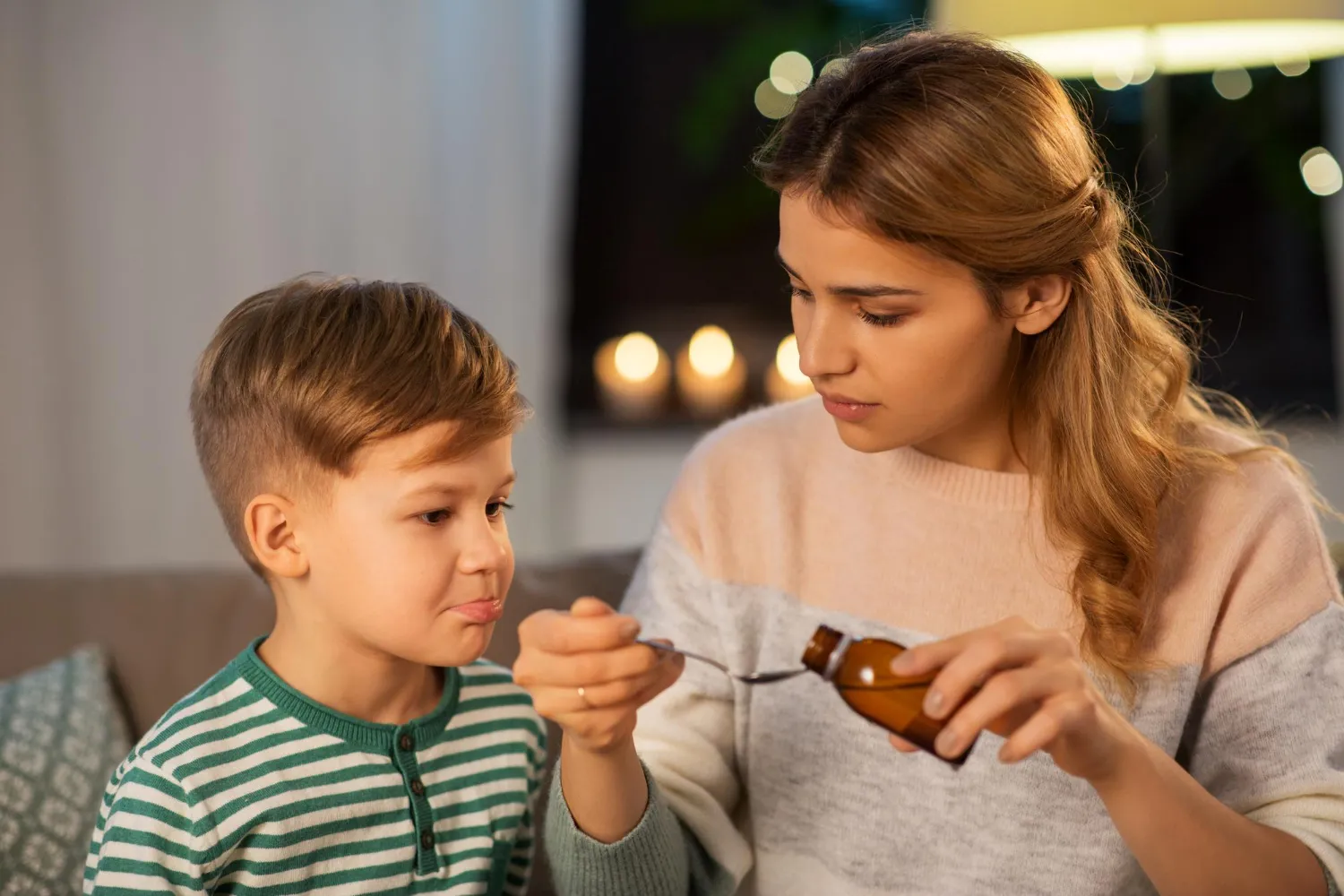 Woman feeds a young boy a spoonful from a bottle, sitting on a couch at night.